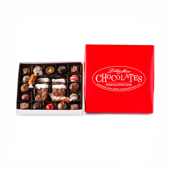 An open box of assorted chocolates size medium displayed next to its red branded lid on a white background. The chocolates include various shapes, sizes, and toppings in shades of brown, milk chocolate, and white including caramels, chocolate covered caramels, truffles, and TorTush our version of a turtle.