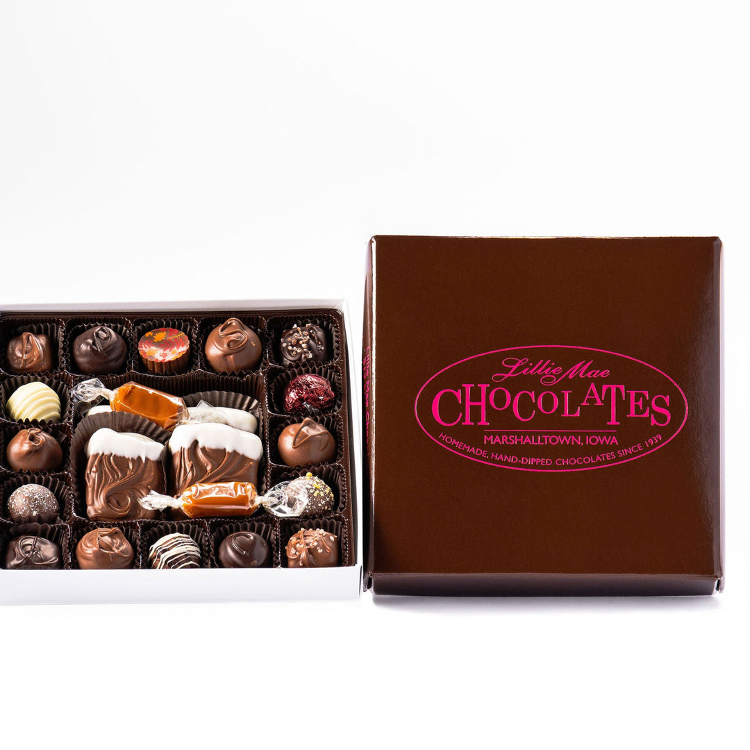 An open box of assorted chocolates displayed next to its brown branded lid on a white background. The chocolates include various shapes, sizes, and toppings in shades of brown, milk chocolate, and white including caramels, chocolate covered caramels, truffles, and TorTush our version of a turtle.