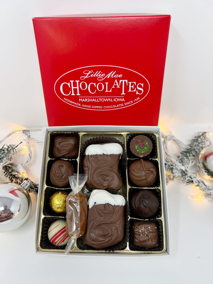 An open box of assorted chocolates displayed next to its red branded lid on a white background. The chocolates include various shapes, sizes, and toppings in shades of brown, milk chocolate, and white including caramels, chocolate covered caramels, truffles, and TorTush our version of a turtle.