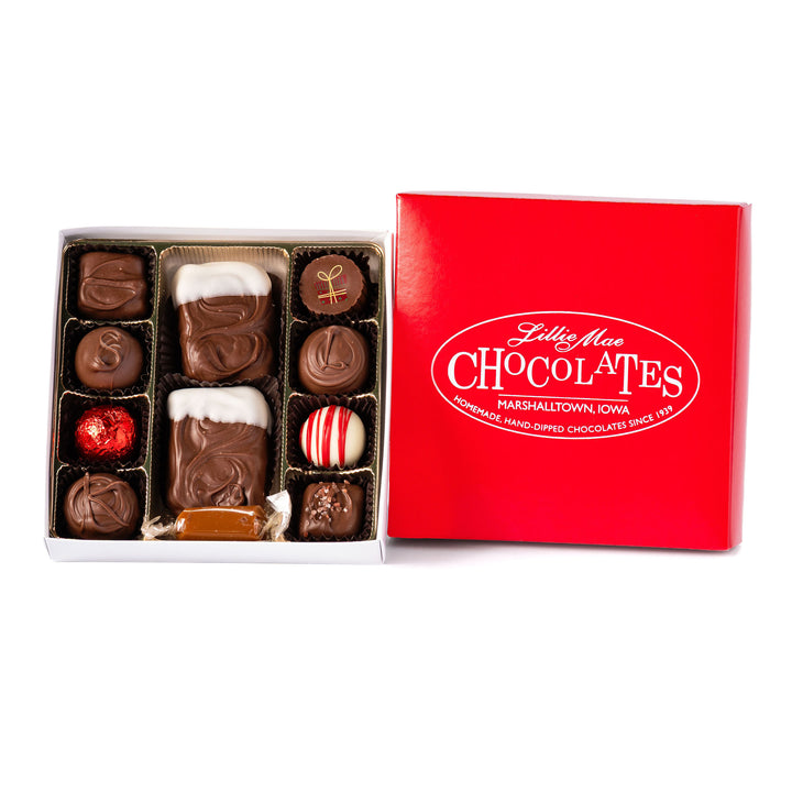 An open box of assorted chocolates size medium displayed next to its red branded lid on a white background. The chocolates include various shapes, sizes, and toppings in shades of brown, milk chocolate, and white including caramels, chocolate covered caramels, truffles, and TorTush our version of a turtle