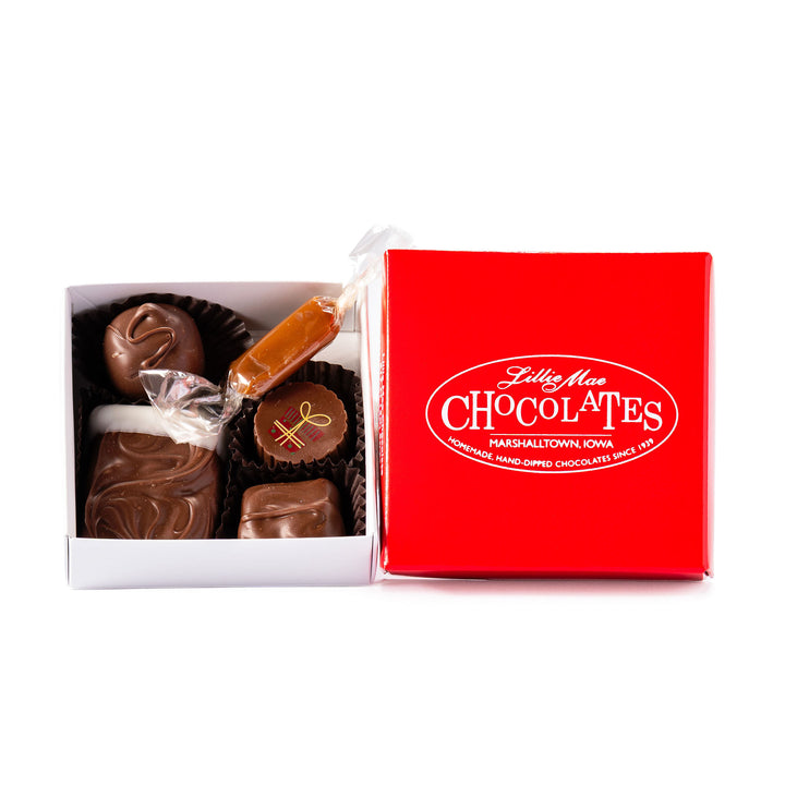An open box of assorted chocolates displayed next to its red branded lid on a white background. The chocolates include various shapes, sizes, and toppings in shades of brown, milk chocolate, and white including caramels, chocolate covered caramels, truffles, and TorTush our version of a turtle