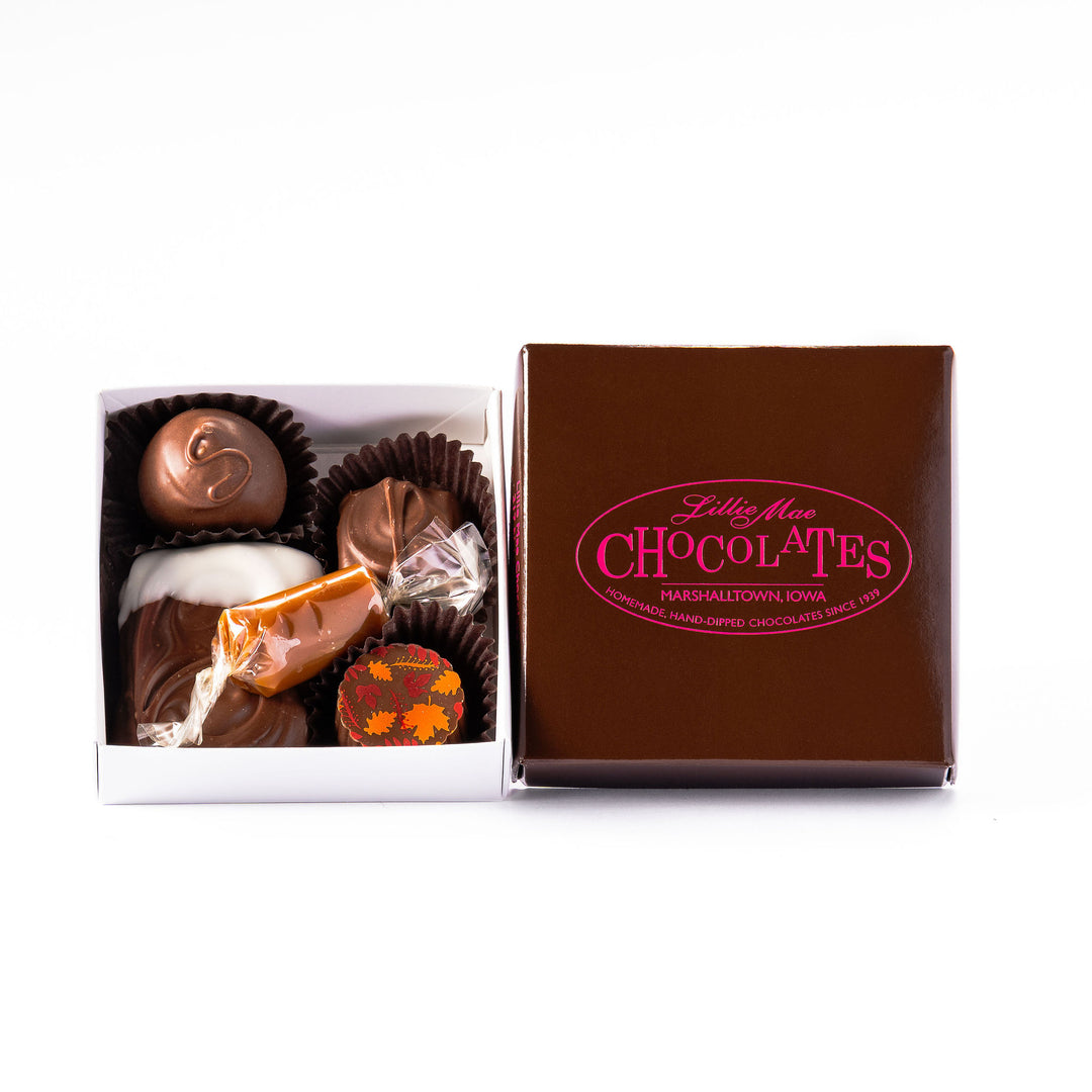 An small open box of assorted chocolates displayed next to its brown branded lid on a white background. The chocolates include various shapes, sizes, and toppings in shades of brown, milk chocolate, and white including caramels, chocolate covered caramels, truffles, and TorTush our version of a turtle.