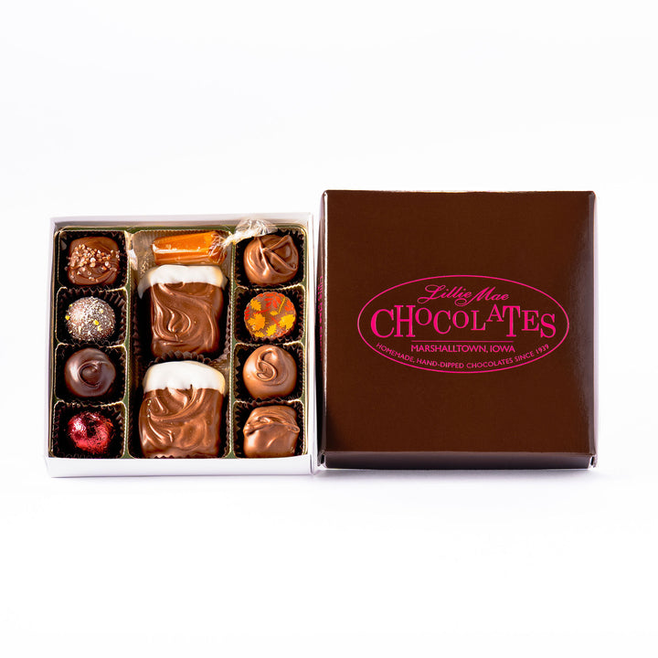 An open box of assorted chocolates size medium displayed next to its brown branded lid on a white background. The chocolates include various shapes, sizes, and toppings in shades of brown, milk chocolate, and white including caramels, chocolate covered caramels, truffles, and TorTush our version of a turtle.