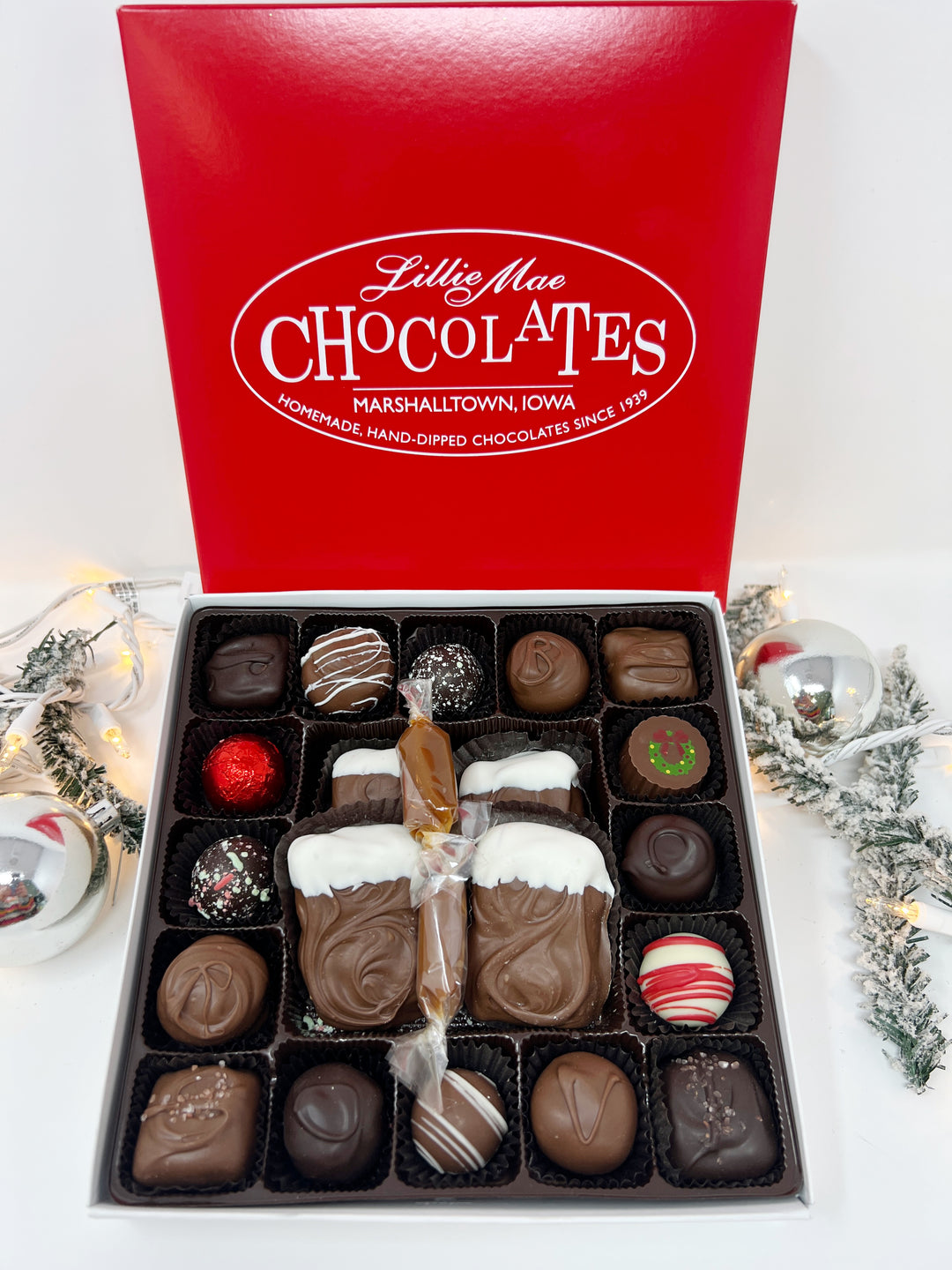 An open box of assorted chocolates displayed next to its red branded lid on a with a holiday background. The chocolates include various shapes, sizes, and toppings in shades of brown, milk chocolate, and white including caramels, chocolate covered caramels, truffles, and TorTush our version of a turtle.