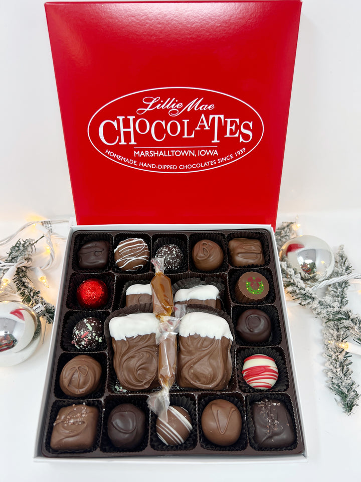 An open box of assorted chocolates displayed next to its red branded lid on a with a holiday background. The chocolates include various shapes, sizes, and toppings in shades of brown, milk chocolate, and white including caramels, chocolate covered caramels, truffles, and TorTush our version of a turtle.