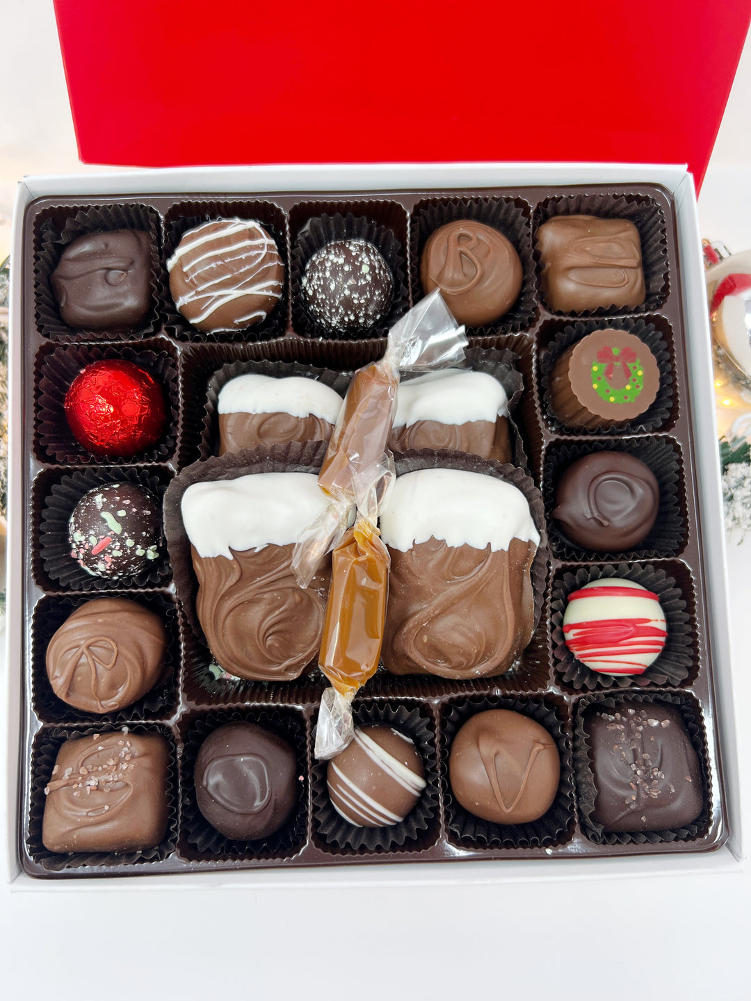 A close up open box of assorted chocolates size large displayed next to its red branded lid on a white background. The chocolates include various shapes, sizes, and toppings in shades of brown, milk chocolate, and white including caramels, chocolate covered caramels, truffles, and TorTush our version of a turtle