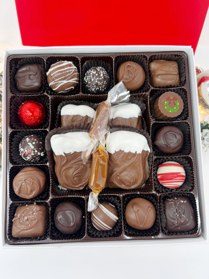 A close up open box of assorted chocolates size large displayed next to its red branded lid on a white background. The chocolates include various shapes, sizes, and toppings in shades of brown, milk chocolate, and white including caramels, chocolate covered caramels, truffles, and TorTush our version of a turtle